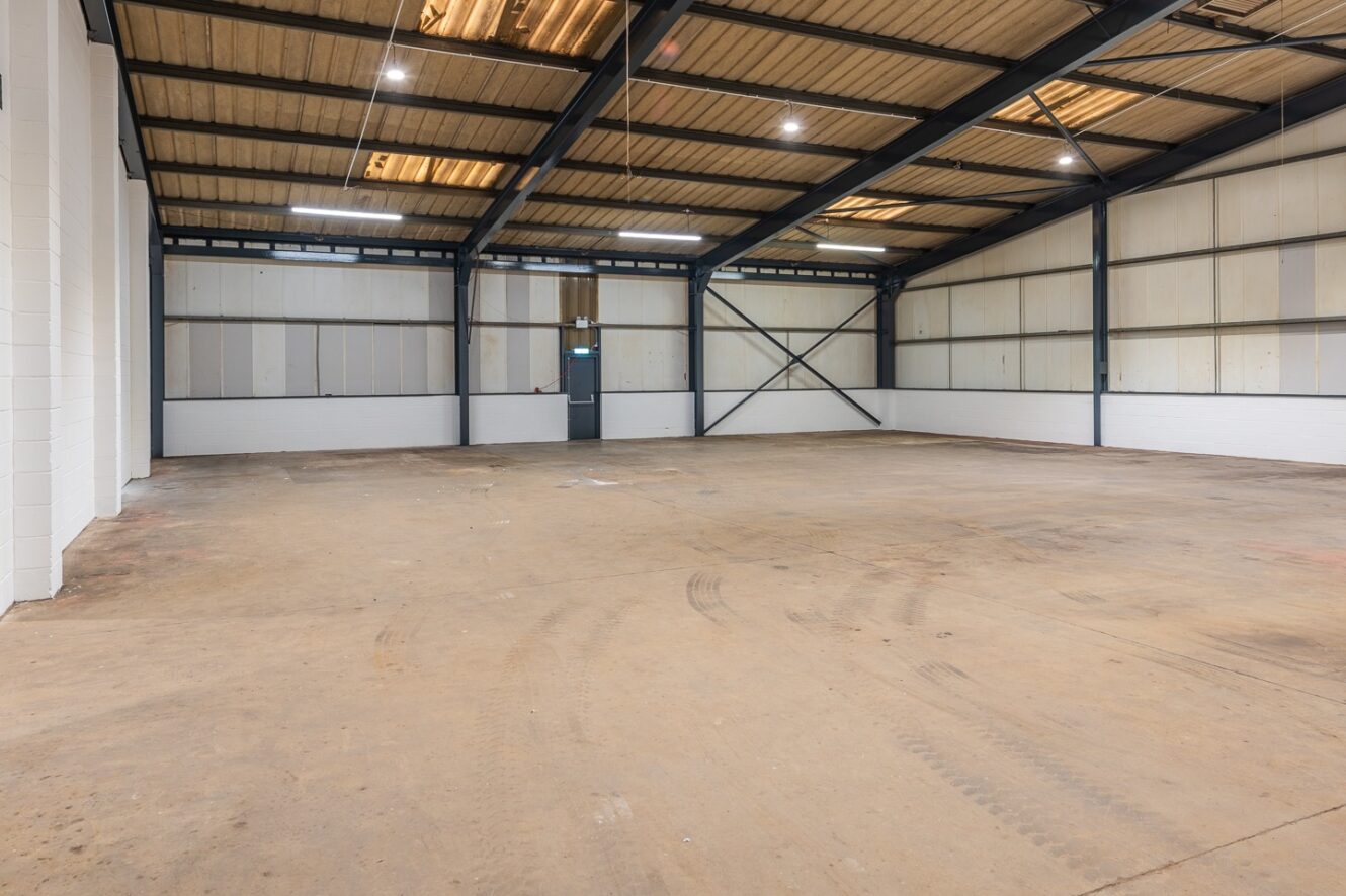 Empty industrial warehouse interior with concrete floor, metal beams, high ceiling, and fluorescent lighting. Door and window visible at the far end. No furniture or equipment present.