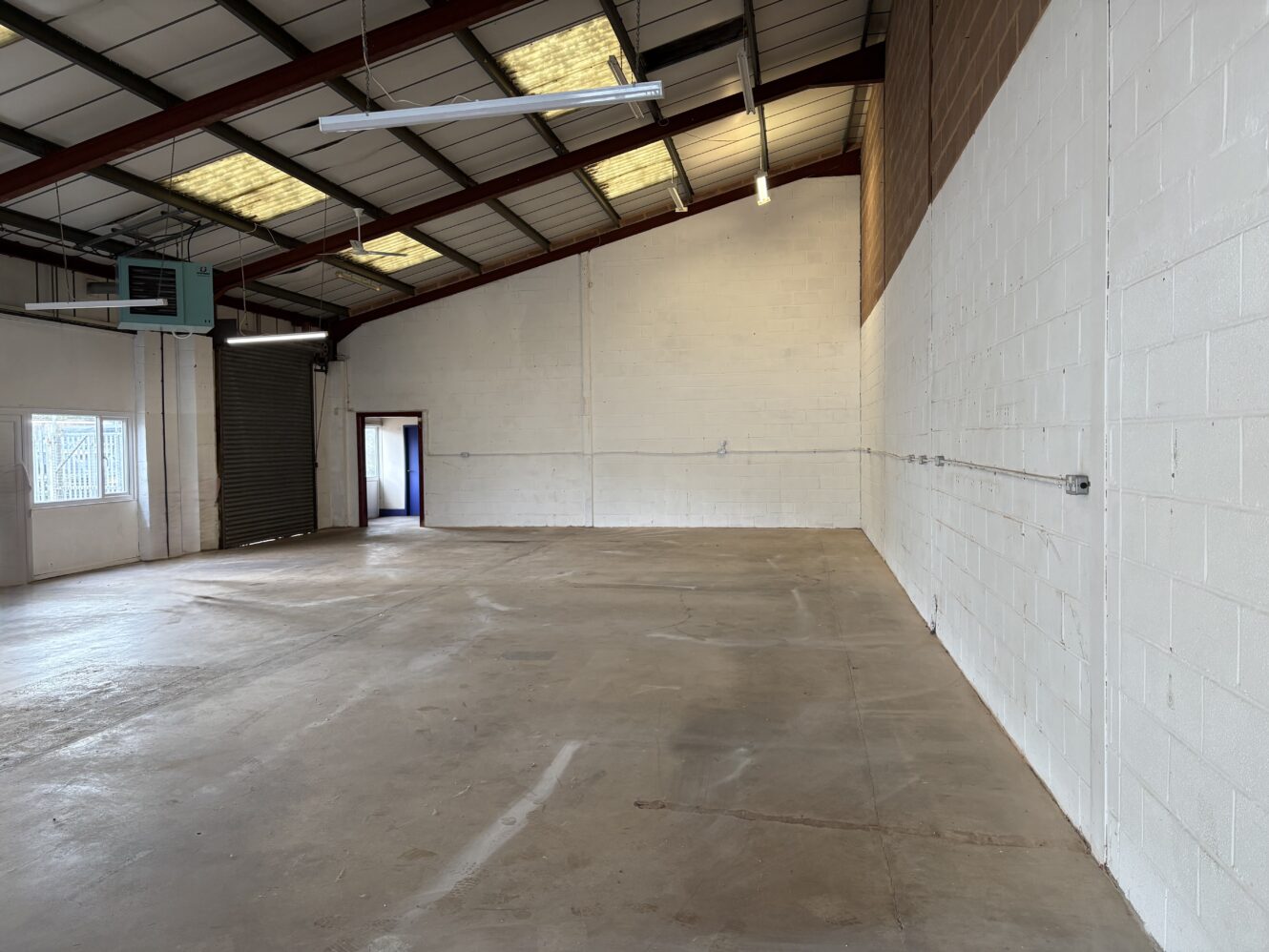 Empty industrial warehouse space with concrete floors, white brick walls, exposed ceiling beams, fluorescent lights, and a large open roller shutter to the left.