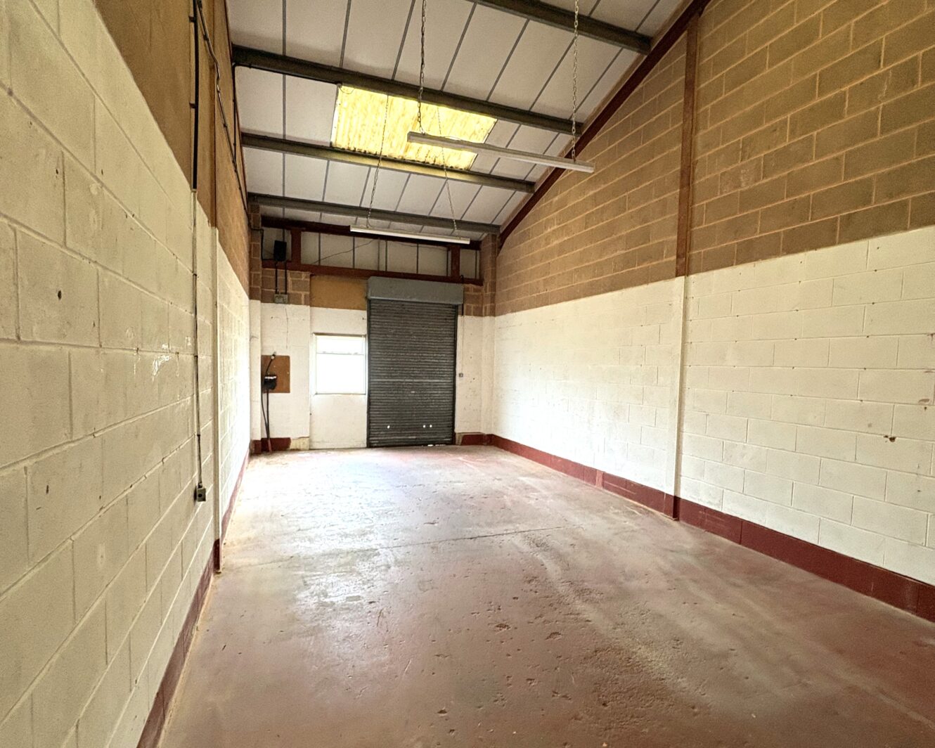 Empty industrial unit with a concrete floor, brick and painted walls, high ceiling, fluorescent lights, and a closed metal roller shutter at the far end.