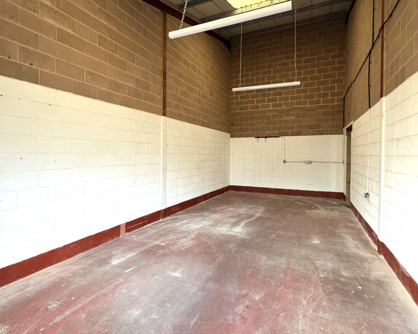Empty industrial room with bare brick and painted white walls, high ceiling, exposed lighting fittings, and a red concrete floor.