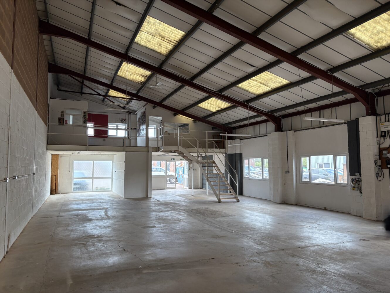Interior of an empty industrial warehouse with white walls, large windows, high ceiling, and a mezzanine level with stairs.