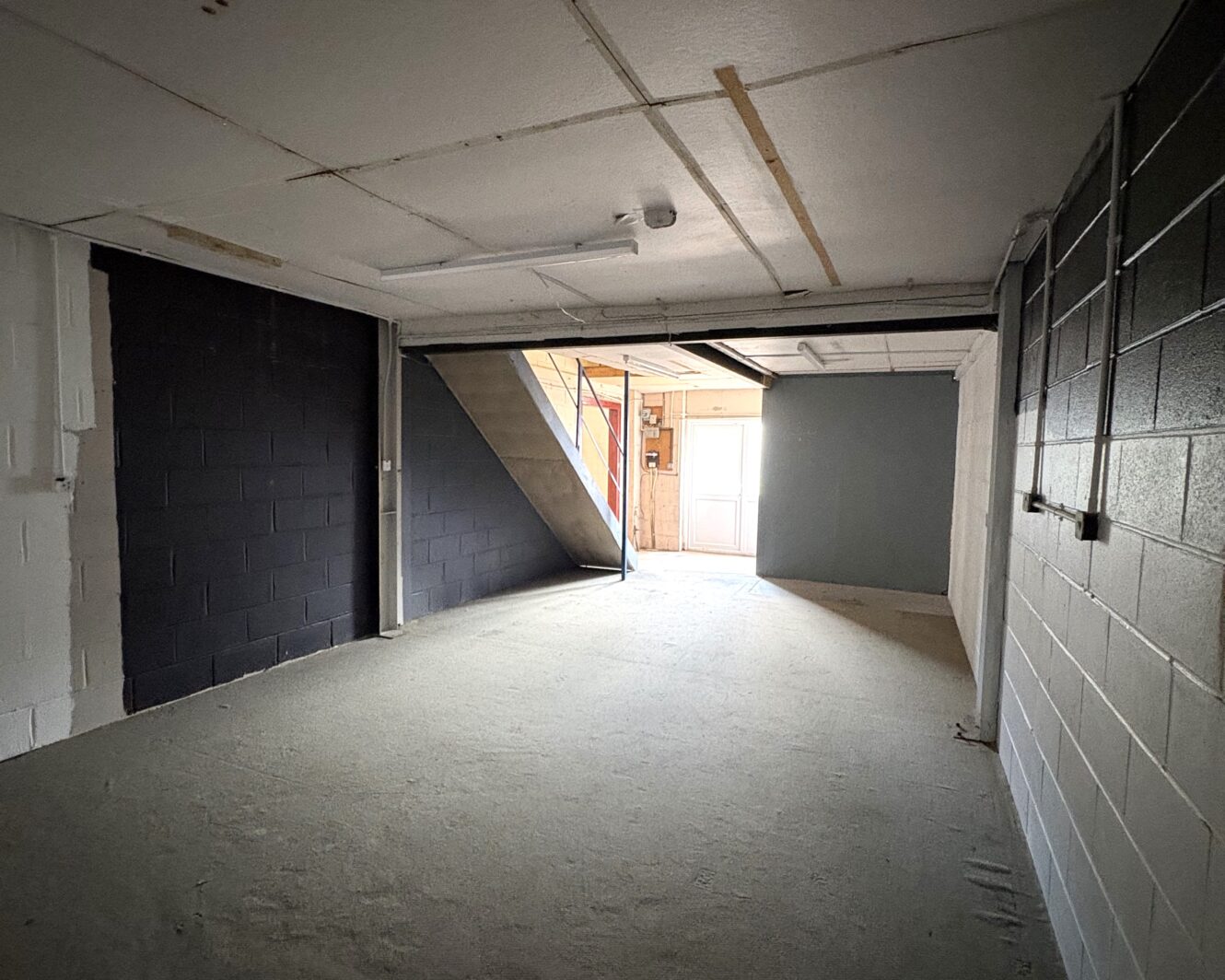 Empty industrial room with a concrete floor, painted breeze block walls, an exposed ceiling, staircase on the left, and an open door at the far end allowing natural light to stream in.
