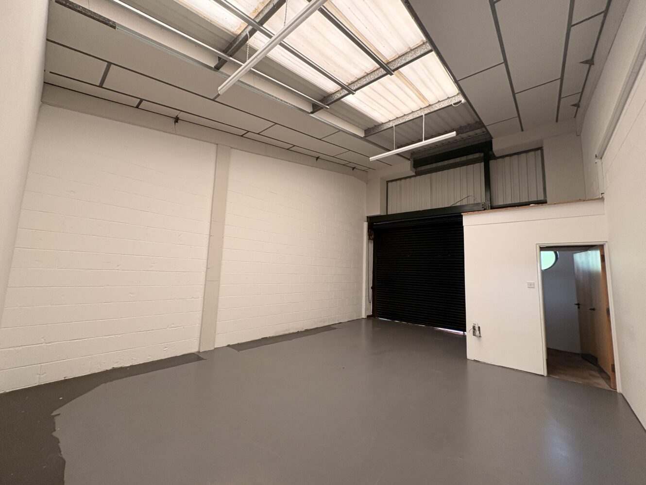 Empty industrial unit with grey painted floor, white brick walls, skylight ceiling panels, and a large black roller shutter door. A small side room with a round window is visible on the right.