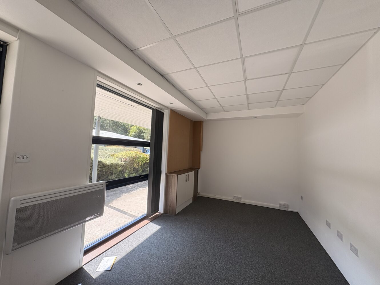 Empty office room with carpeted floor, large window, wall heater, built-in cupboard, and white ceiling tiles. Sunlight streams in through the window, illuminating part of the floor.