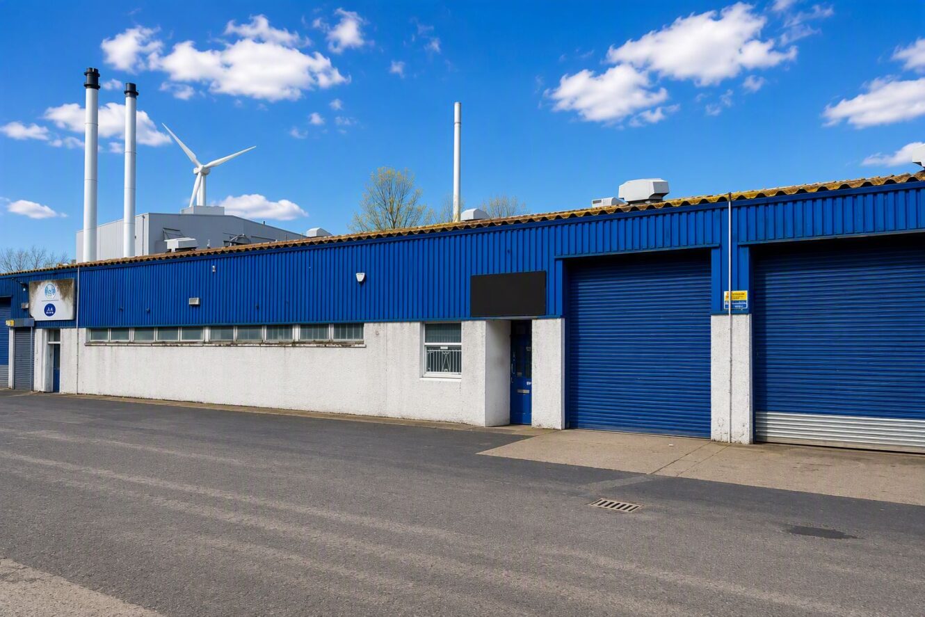 Blue industrial warehouse with closed roller shutters, white lower walls, and chimney stacks; a wind turbine is visible in the background under a blue sky with scattered clouds.