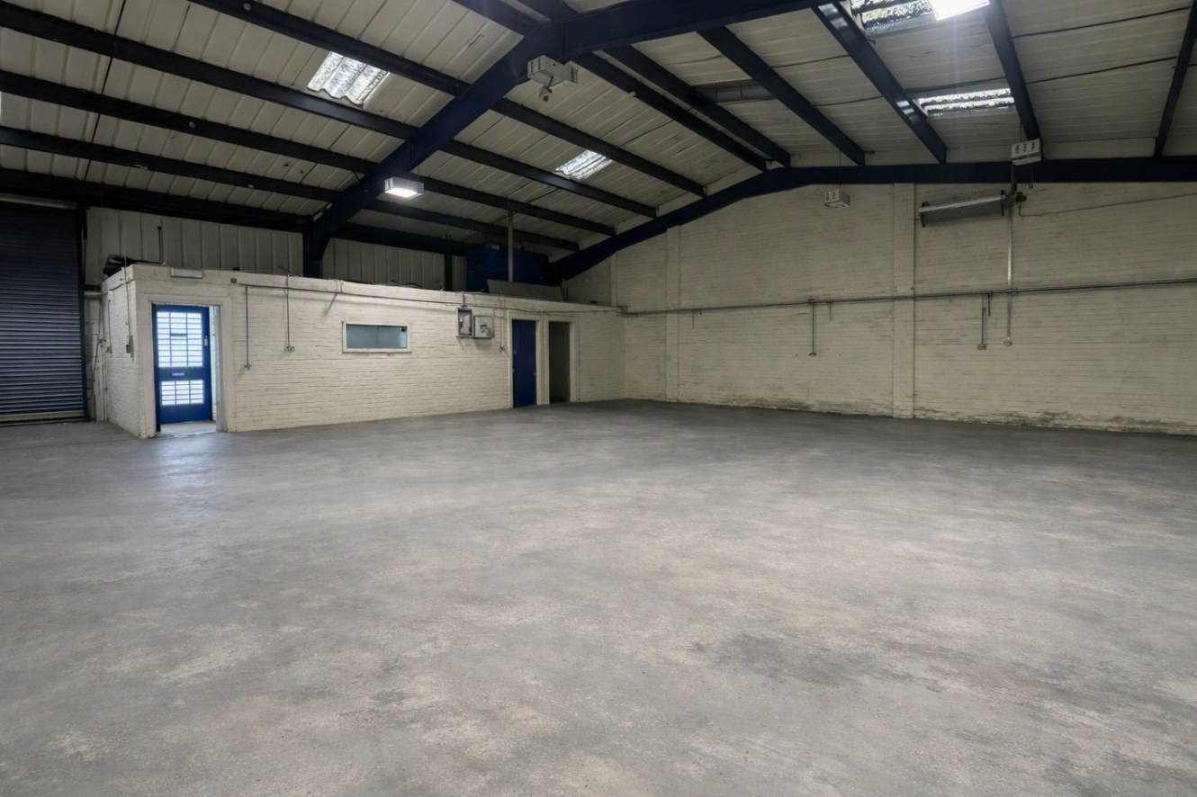 Large empty warehouse with concrete floor, high ceiling, metal beams, white brick walls, fluorescent lights overhead, and blue doors on the left side of the space.
