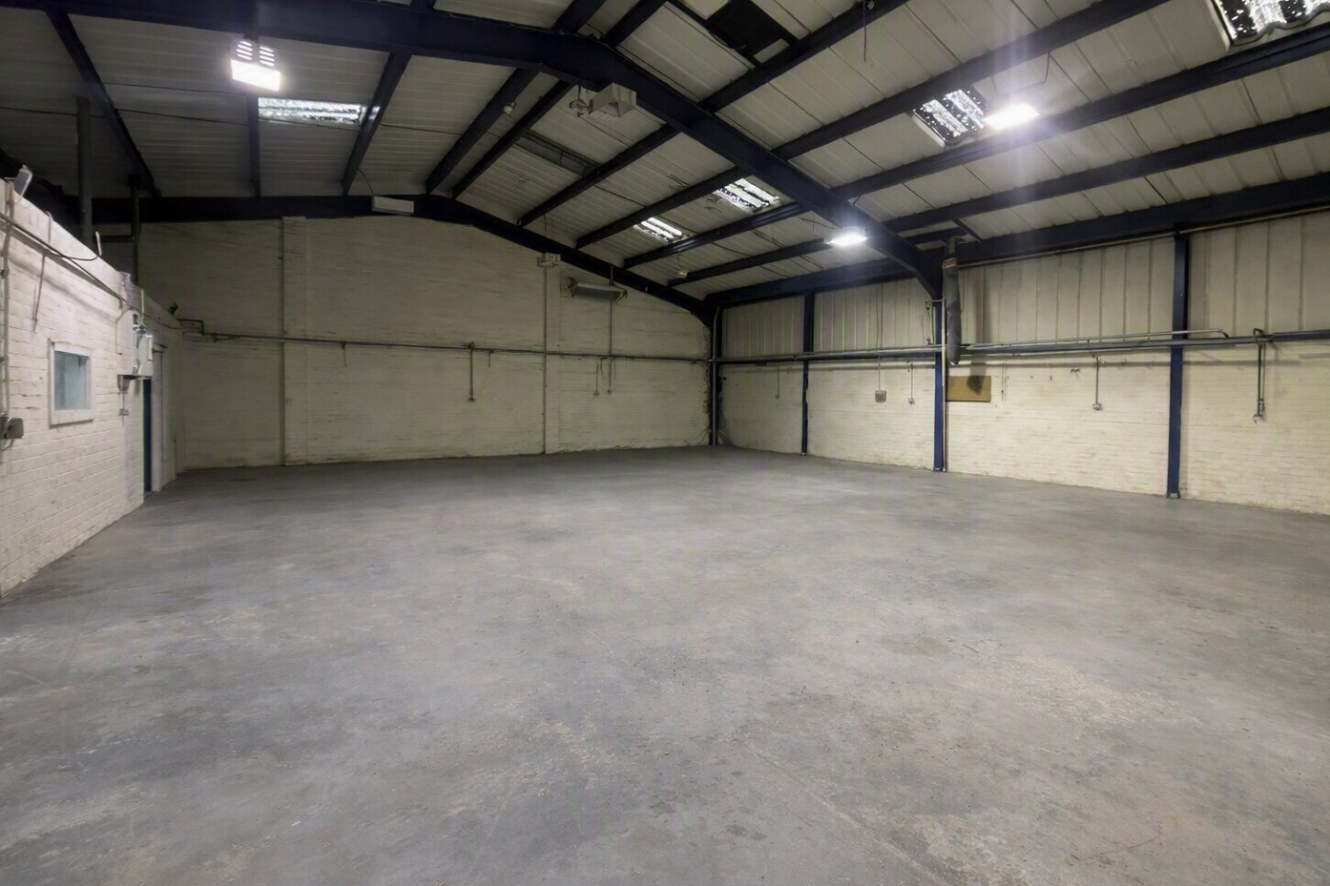 Empty industrial warehouse with concrete floor, white brick walls, and exposed steel beams under a high ceiling with multiple rooflights.