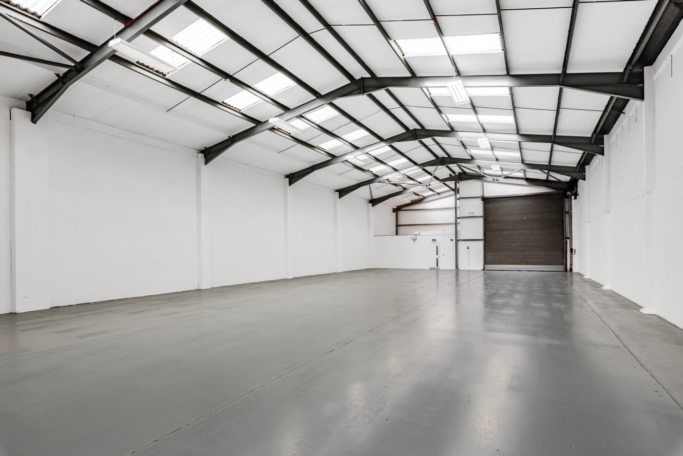 Large, empty industrial warehouse with white walls, high ceiling, exposed metal beams, and a polished concrete floor, illuminated by fluorescent lights.