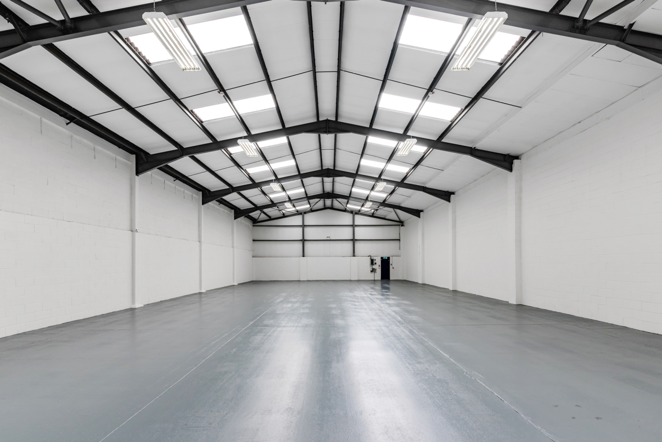 Empty industrial warehouse with white walls, grey polished floor, and overhead fluorescent lighting beneath a vaulted ceiling.
