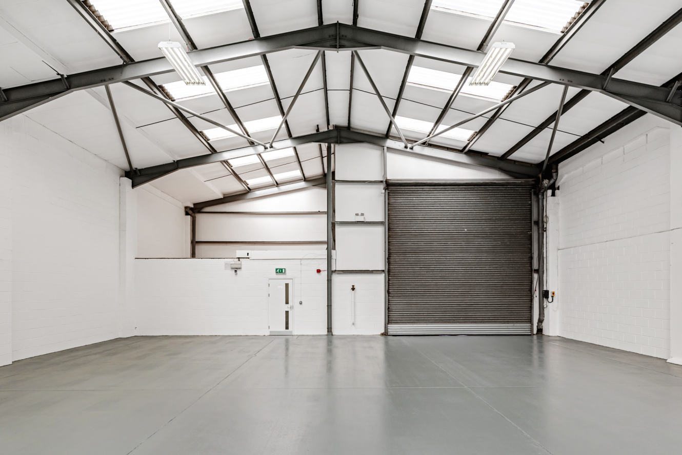 Empty industrial warehouse with white walls, grey concrete floor, metal ceiling beams, skylights, fluorescent lights, a closed roller shutter door, and a single door on the left.