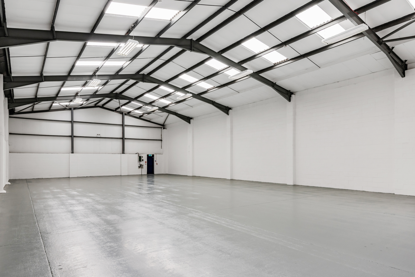 Empty industrial warehouse with white walls, a high ceiling, steel beams, and a grey concrete floor. Fluorescent lights and skylights provide bright, even illumination throughout the space.