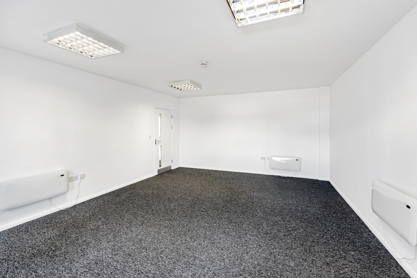 Empty white office room with grey carpet, fluorescent ceiling lights, wall heaters, and a closed door.