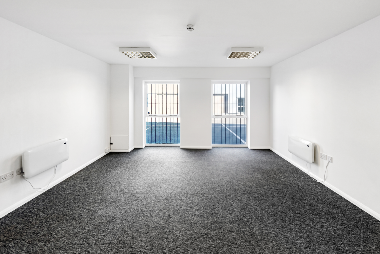 Empty white office room with grey carpet, two ceiling lights, two electric heaters, and a window with vertical bars letting in daylight.
