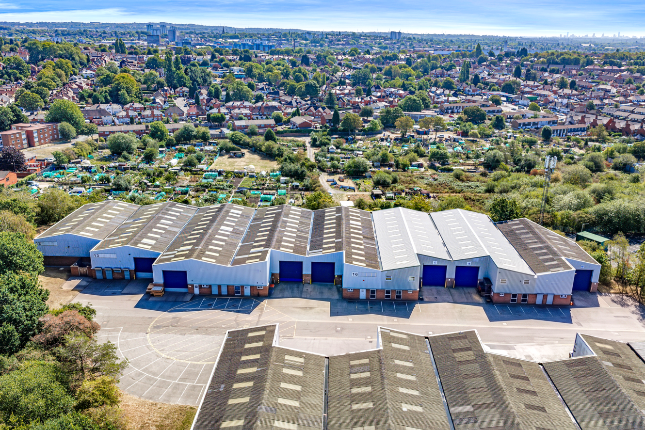 Aerial view of industrial warehouses with blue doors, open car park in front, and residential neighbourhood surrounded by greenery in the background.