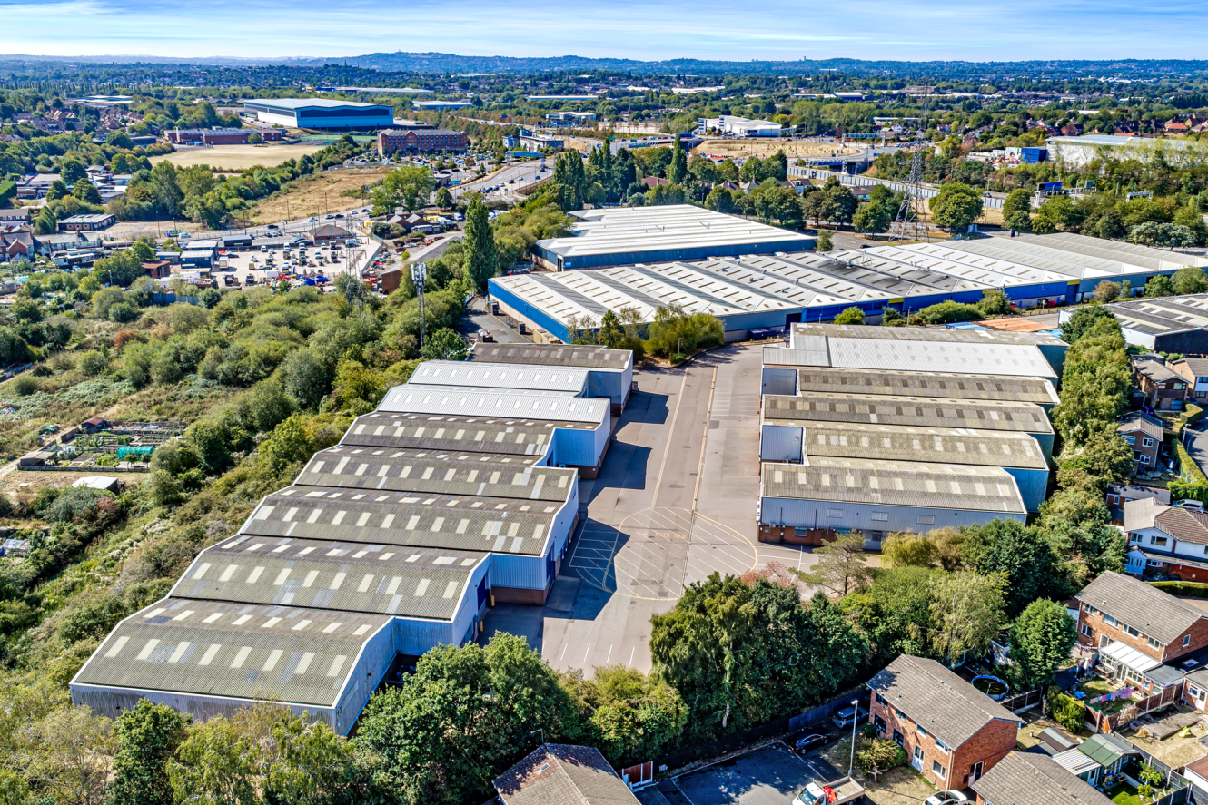 Aerial view of large industrial warehouses with white roofs, surrounded by trees, roads, and nearby residential houses on a clear day.
