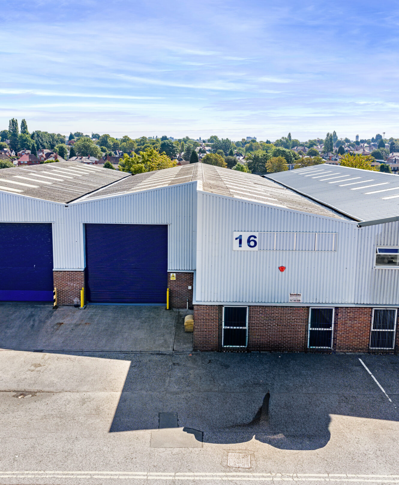 A large industrial warehouse with three blue roller shutters and a sign labelled 16 is shown on a clear day, with trees and houses visible in the background.