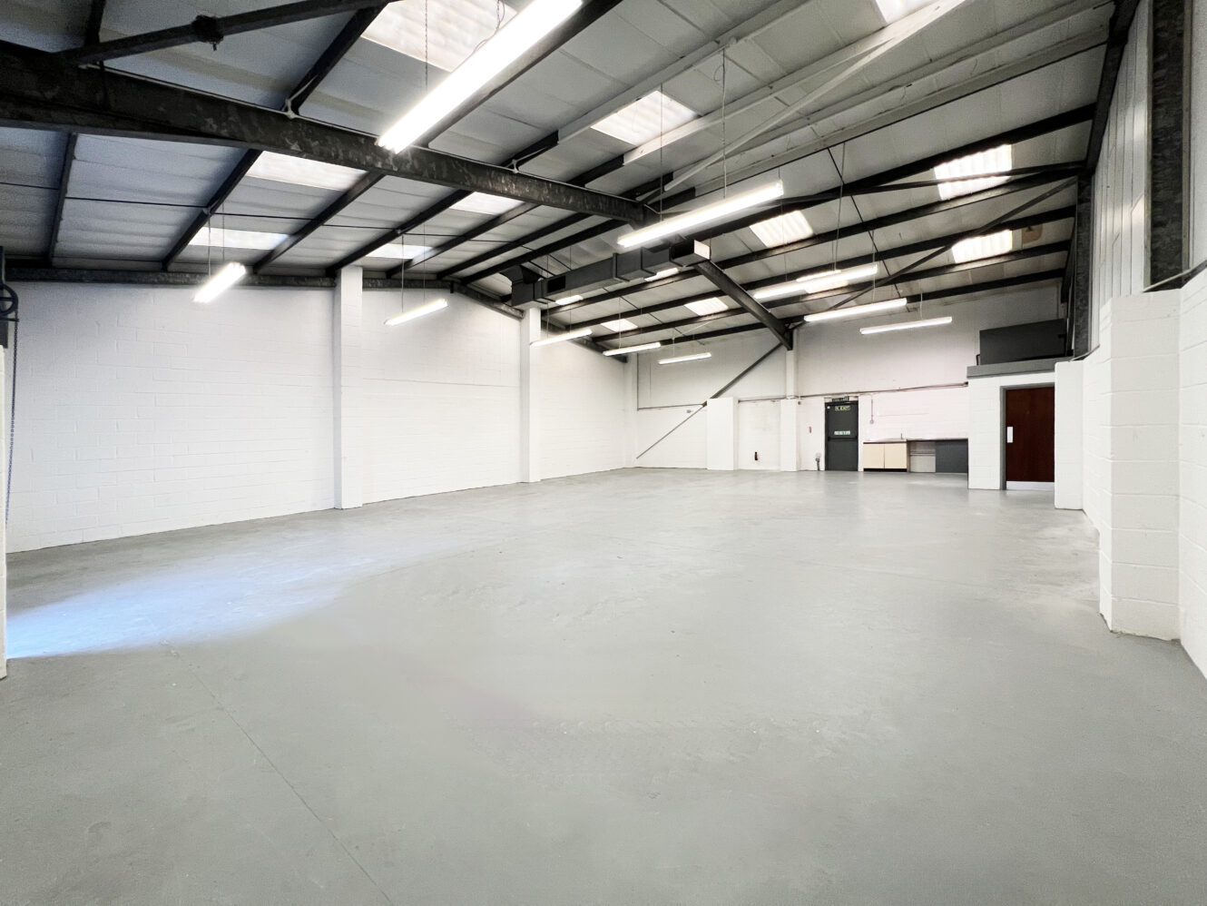 Empty warehouse interior with white walls, concrete floor, exposed steel beams, fluorescent lighting, and a small office area in the back right corner.