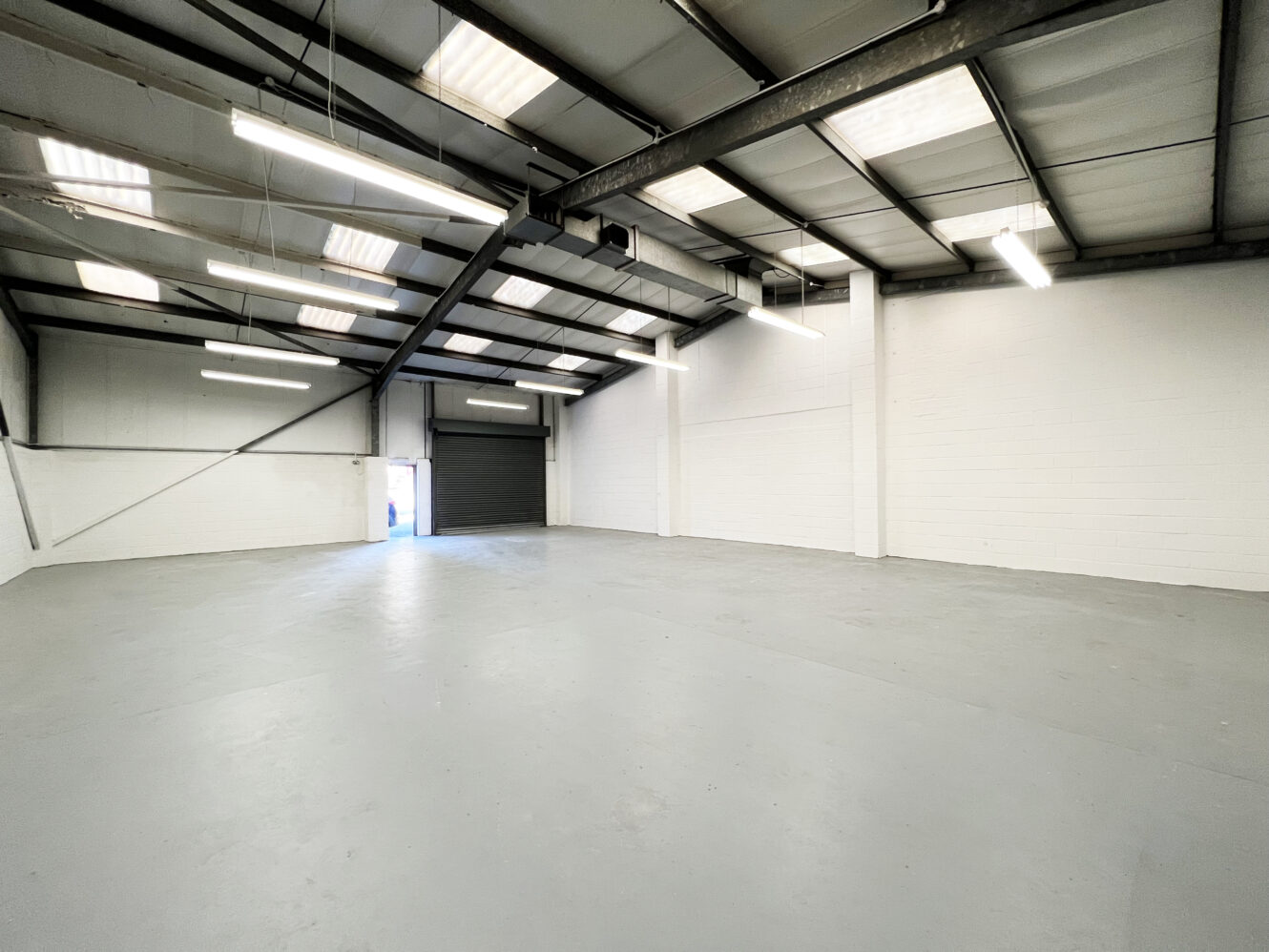 Spacious empty warehouse with white walls, a concrete floor, metal beams, ceiling lights, and a large open garage door letting in daylight.