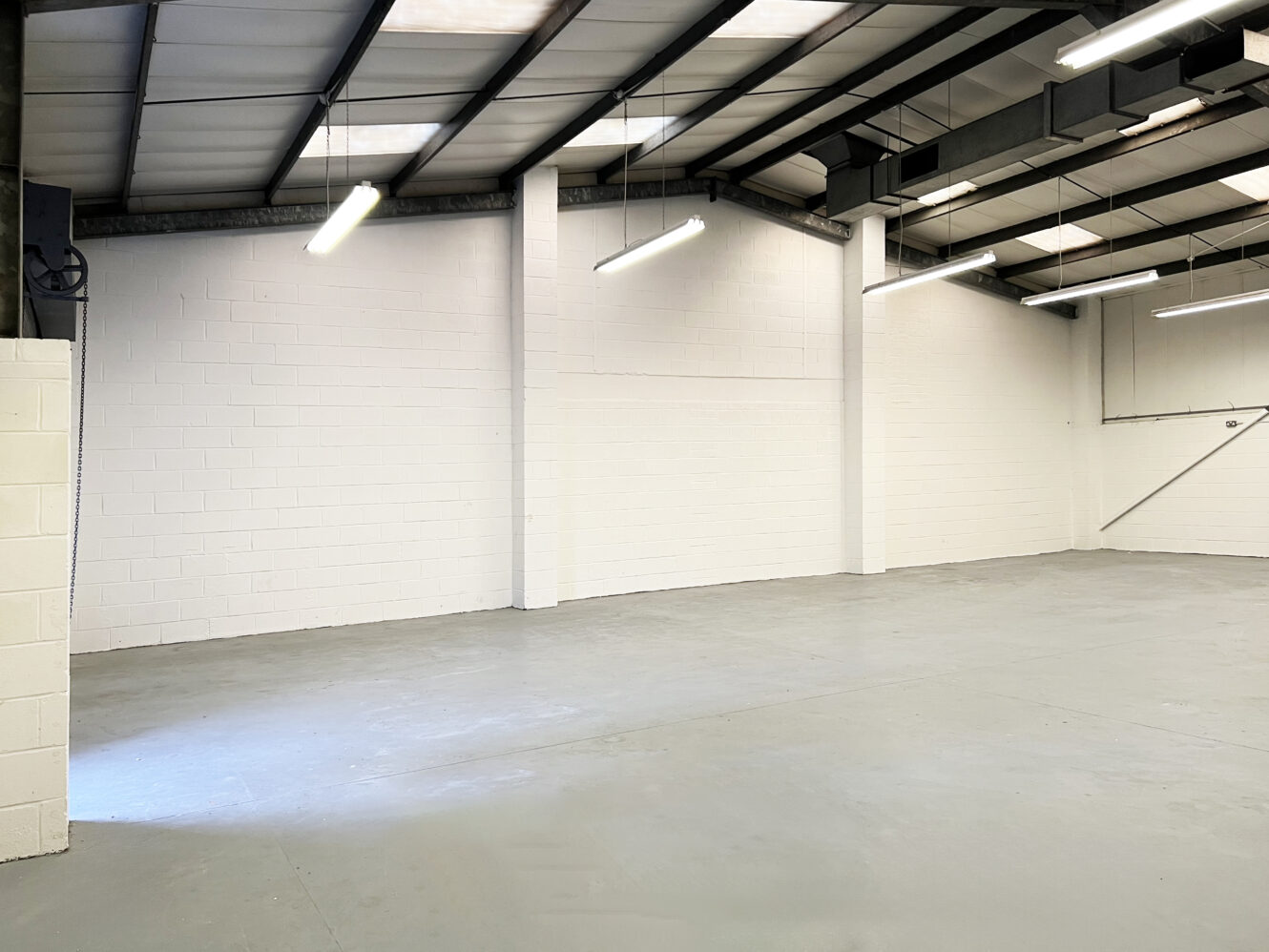 Empty warehouse interior with white painted brick walls, grey concrete floor, exposed beams, and fluorescent ceiling lights.