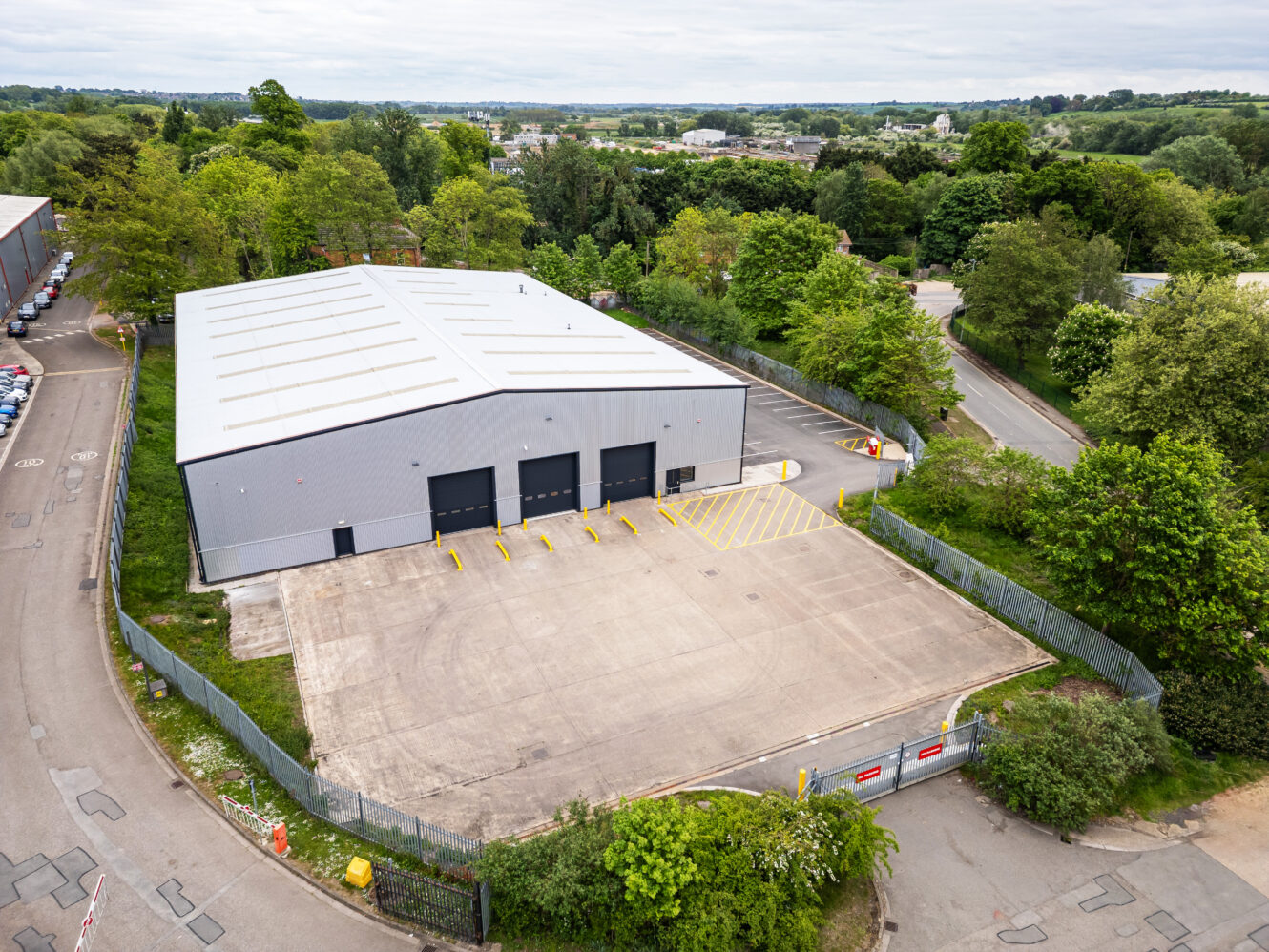 A large grey warehouse with three loading bays and a fenced concrete yard, surrounded by trees and roads, with other buildings and greenery in the background.