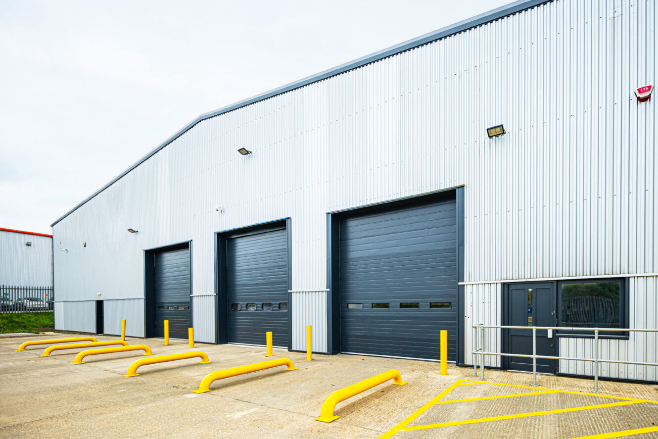 Exterior of a modern industrial warehouse with three large black loading bay doors, yellow safety barriers, and a metal ramp by the entrance.