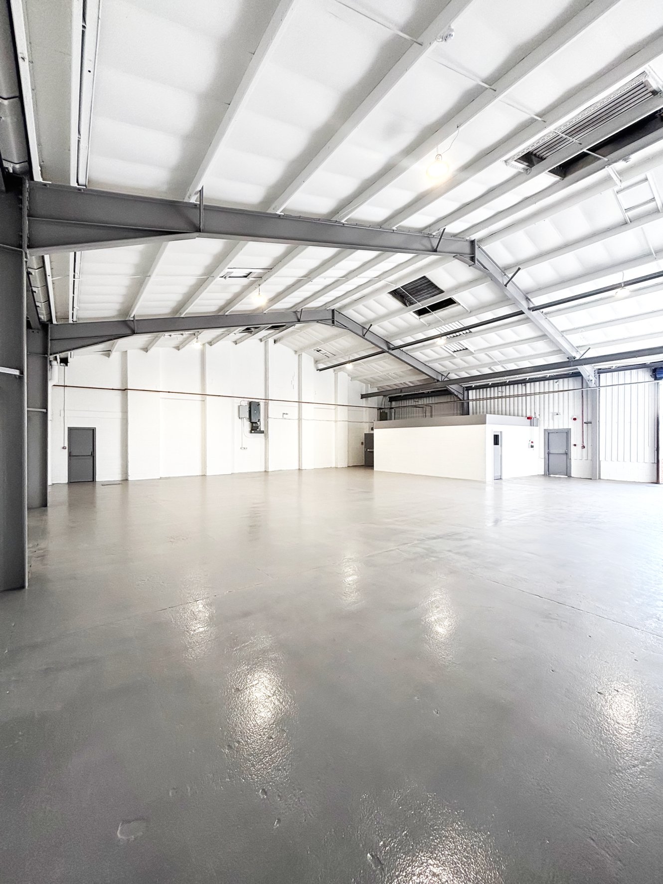 Empty industrial warehouse with white walls, grey polished floor, exposed beams, high ceiling, fluorescent lights, and multiple doors.