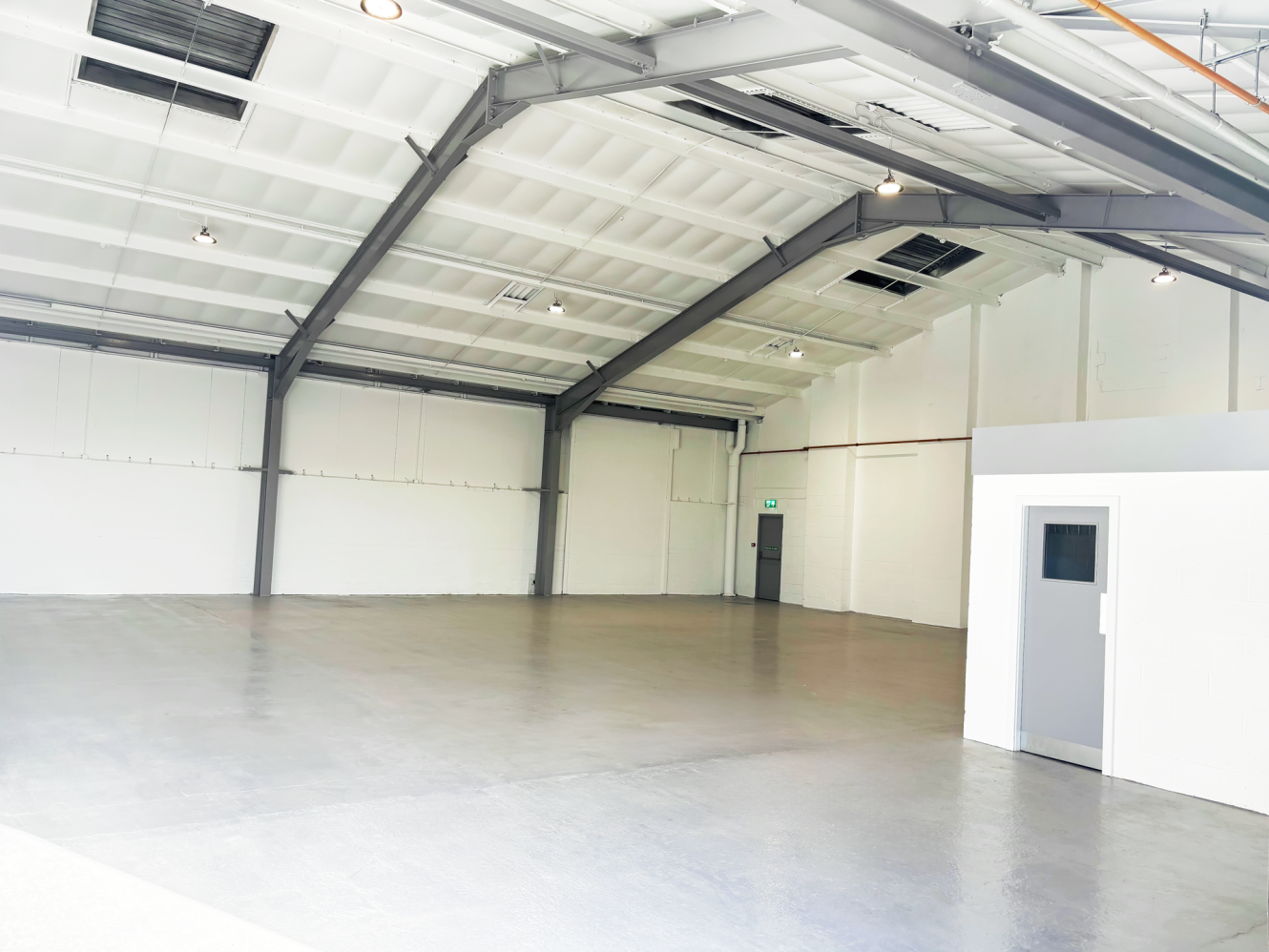 Interior of an empty warehouse with white walls, a grey concrete floor, exposed ceiling beams, and a small enclosed room with a door and window in the corner.