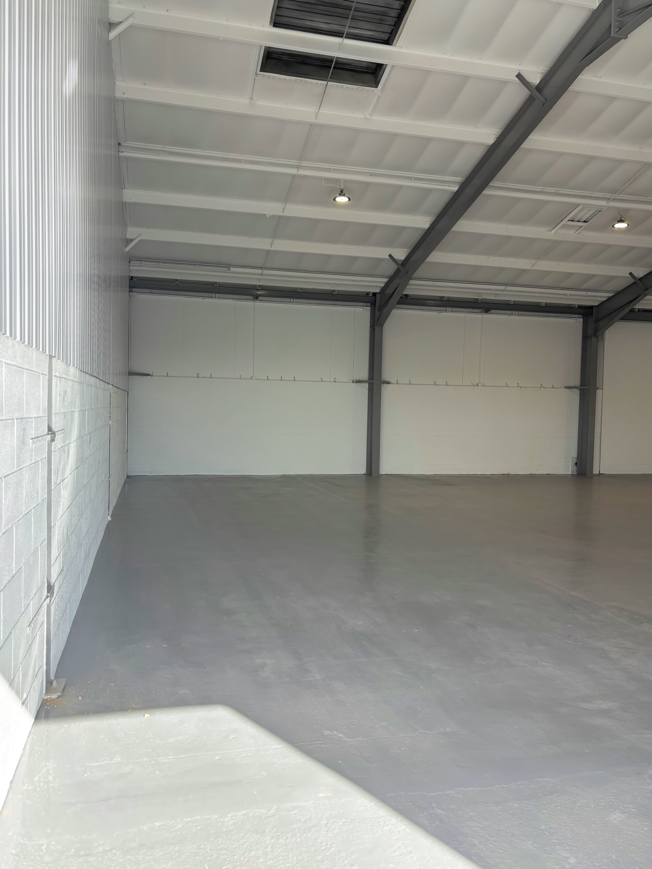 Empty warehouse interior featuring a grey concrete floor, white painted walls, high ceiling, metal beams, and overhead lights.
