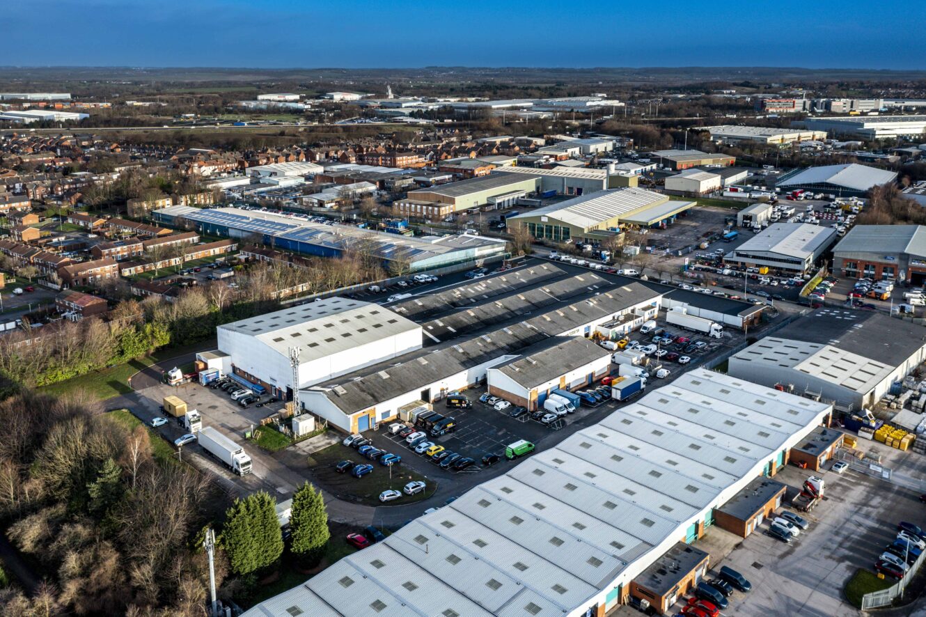 Aerial view of an industrial estate with large warehouses, office buildings, parked vehicles, and nearby residential areas under a clear blue sky.