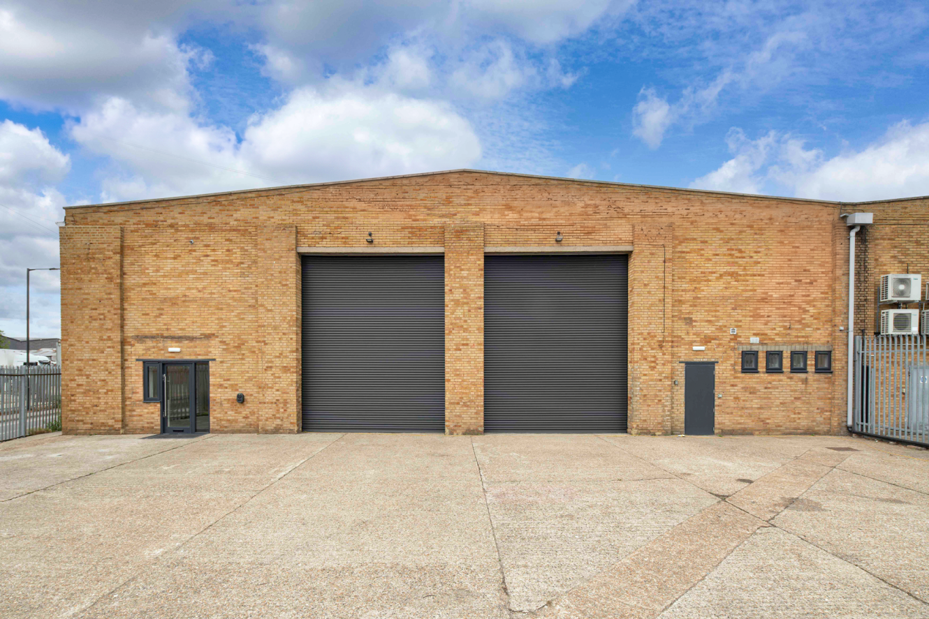 A large brick warehouse with two closed black roller shutters, a small side door, exterior utility boxes, and a wide empty concrete forecourt in front under a partly cloudy sky.