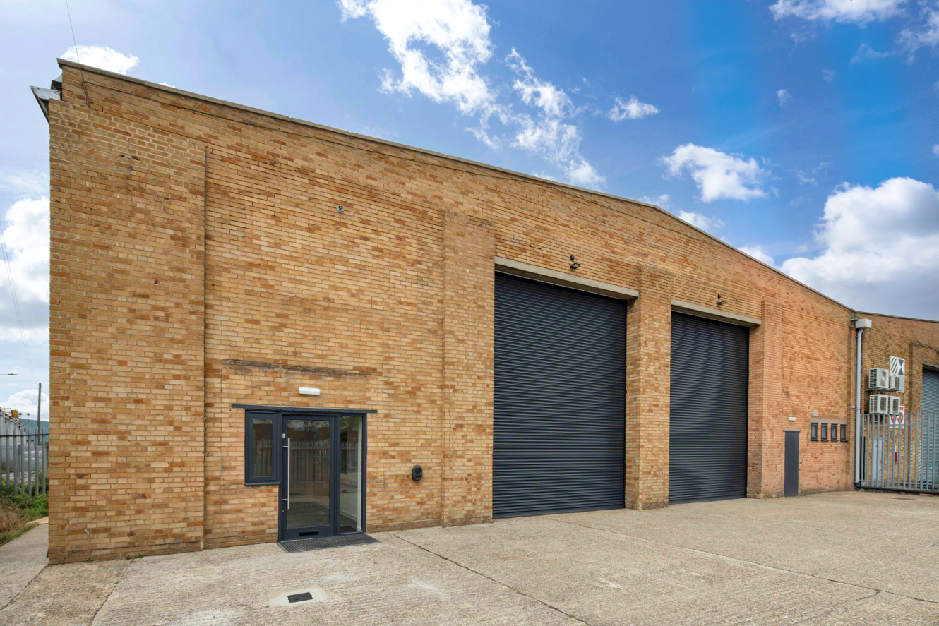 A large brick industrial building with two black roller shutters, a glass entrance door, and a paved forecourt under a partly cloudy sky.