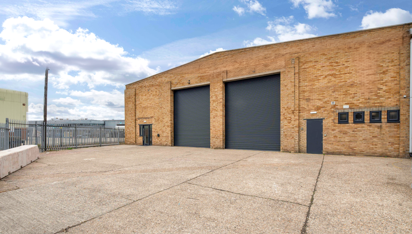 A large industrial building with two closed roller shutters, a smaller entrance door, and a fenced area under a partly cloudy sky.