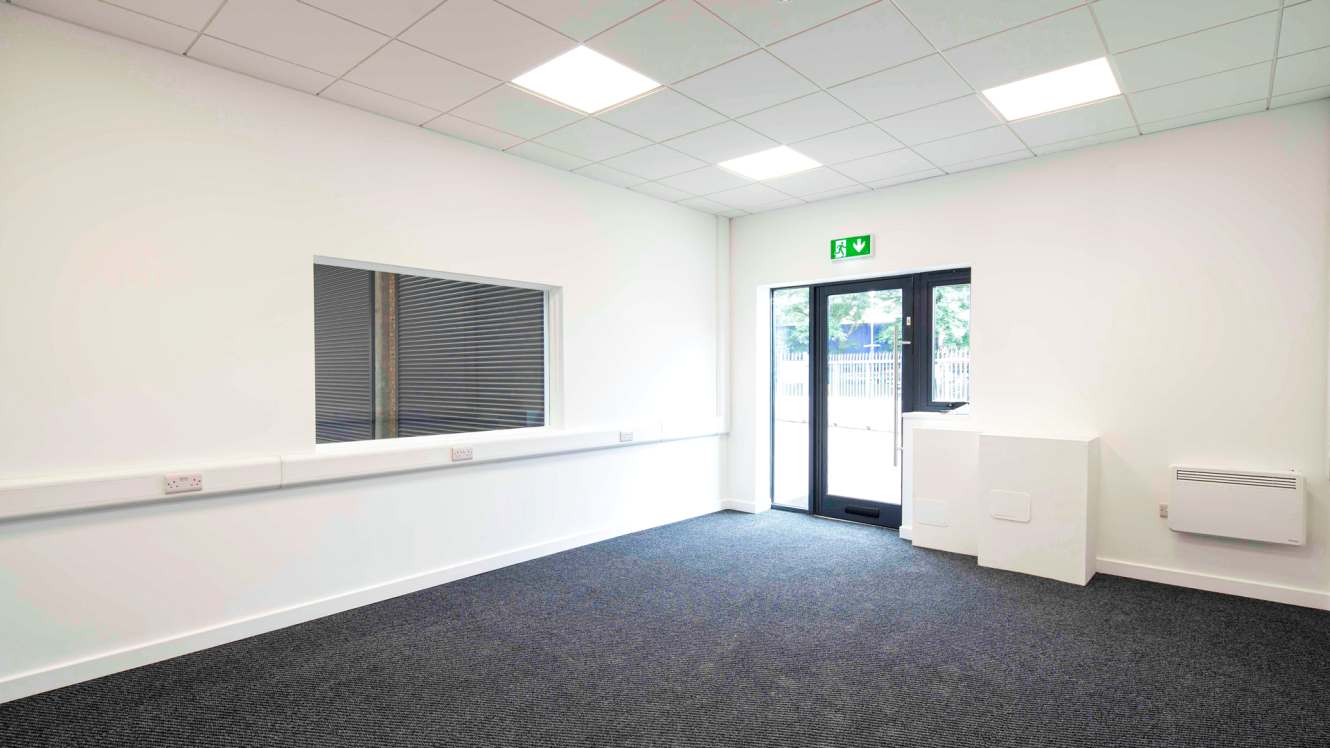 Empty office space with white walls, a carpeted floor, a large window, a glass door displaying an exit sign above it, and a heater mounted on the wall.