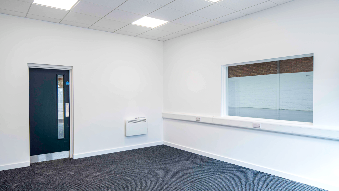 Empty room with white walls, dark carpet, a closed door, a wall heater, plug sockets, and a large internal window showing another empty space.