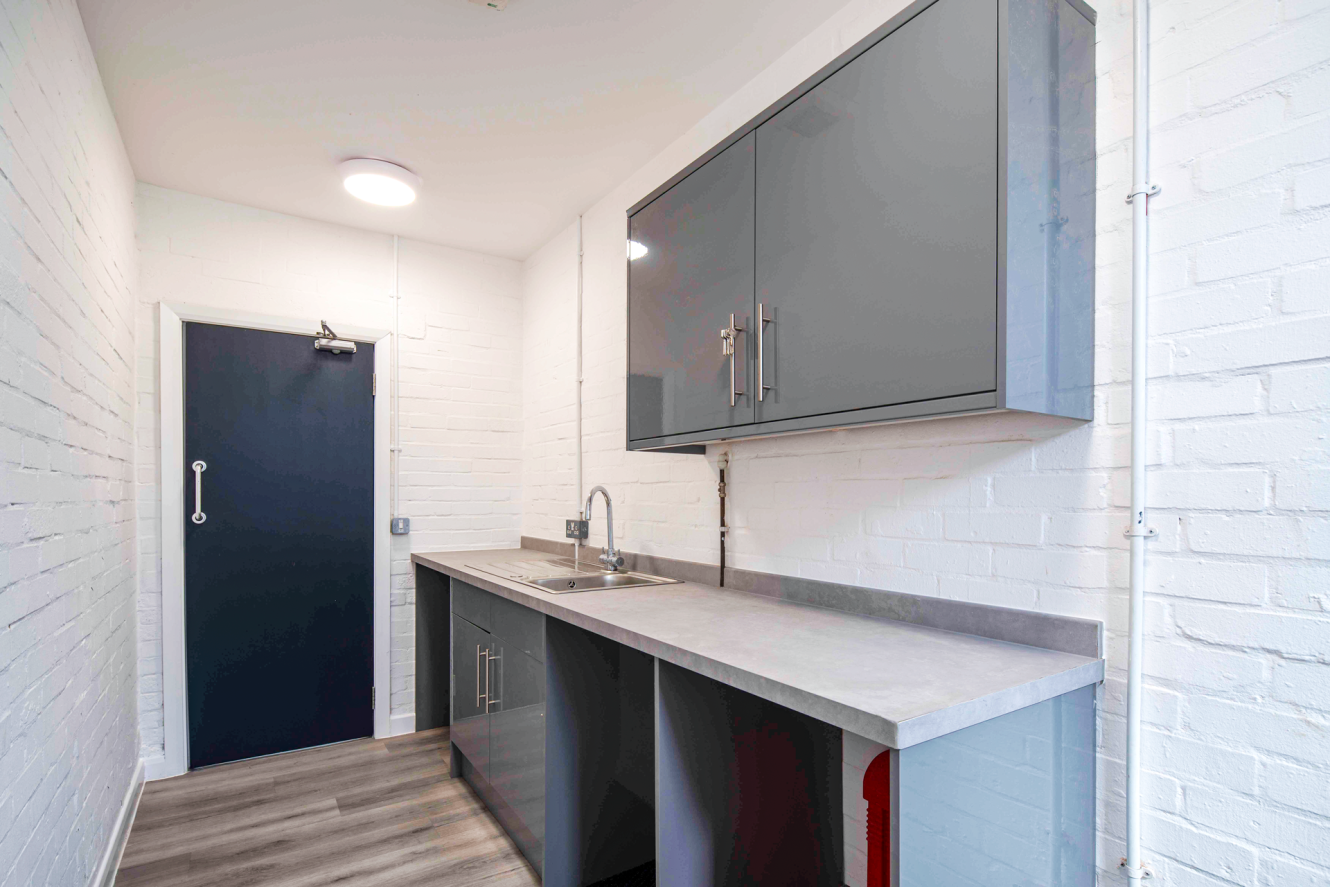 Narrow kitchen with grey cupboards, a stainless steel sink, light wood flooring, and white brick walls.