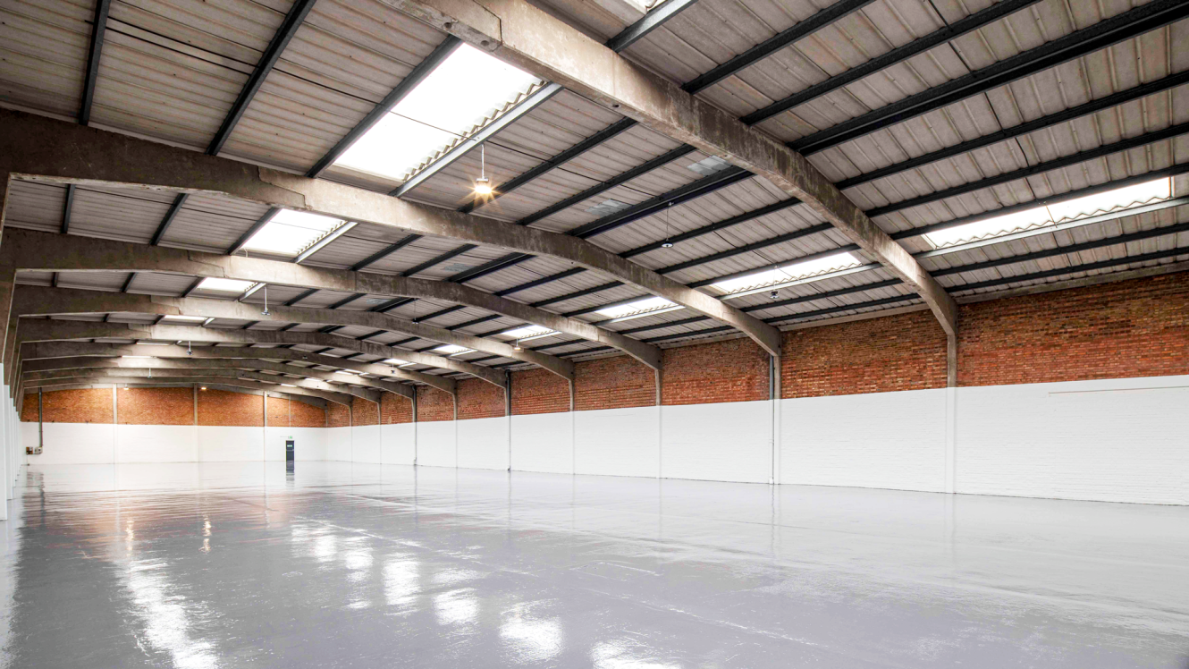 Spacious, empty warehouse with a polished floor, exposed brick walls, and a curved metal roof featuring rooflights.