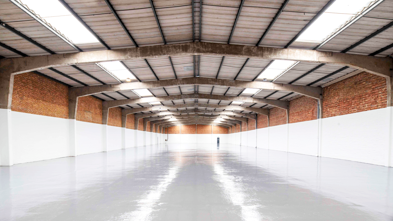 Empty warehouse with brick and white walls, high ceiling, skylights, and polished concrete floor, viewed from the centre looking towards the far end.