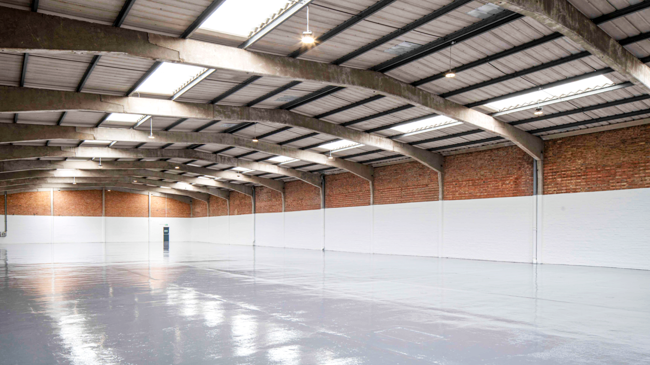 Large, empty warehouse with high arched ceiling, exposed beams, brick upper walls, white lower walls, and a shiny concrete floor.