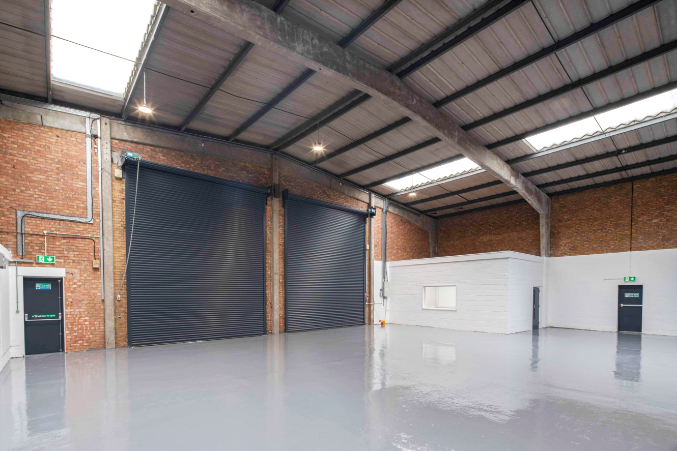 Interior of an empty warehouse with two large black roller shutters, brick walls, polished grey floor, and a small white office structure on the right.
