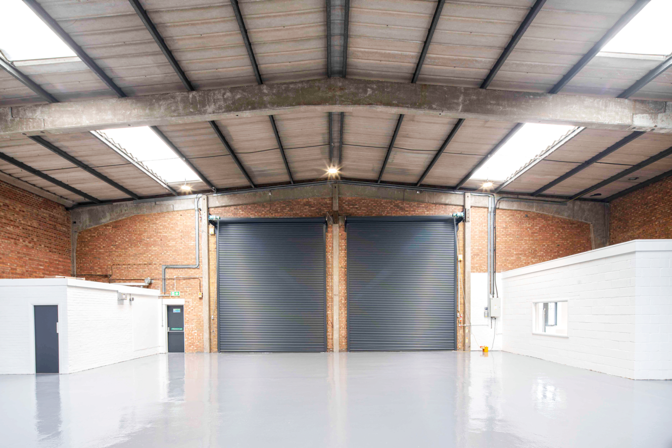 Interior of an empty warehouse featuring brick walls, two large grey roller doors, a polished concrete floor, and small white office rooms situated on both the left and right sides.