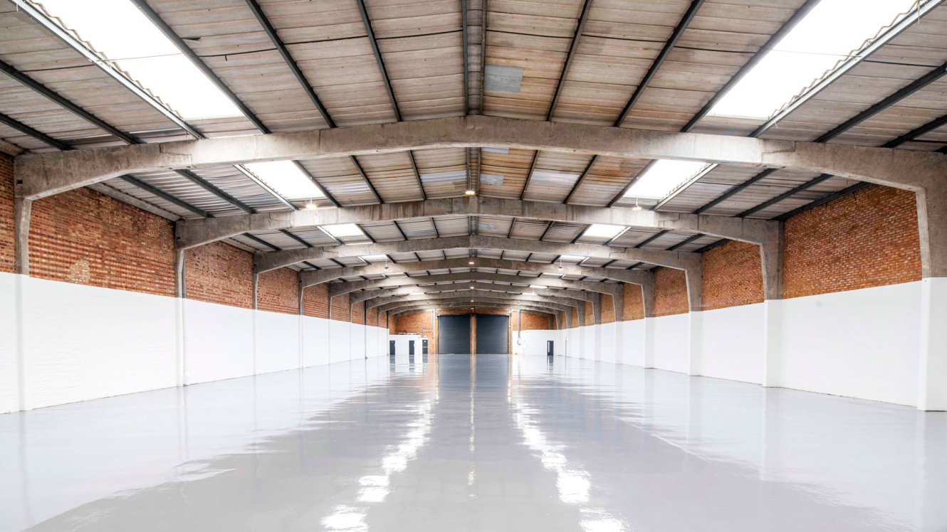 Empty warehouse with polished floor, high ceiling, brick walls, and skylights. Space is well-lit and clean, with a large closed door at the far end.