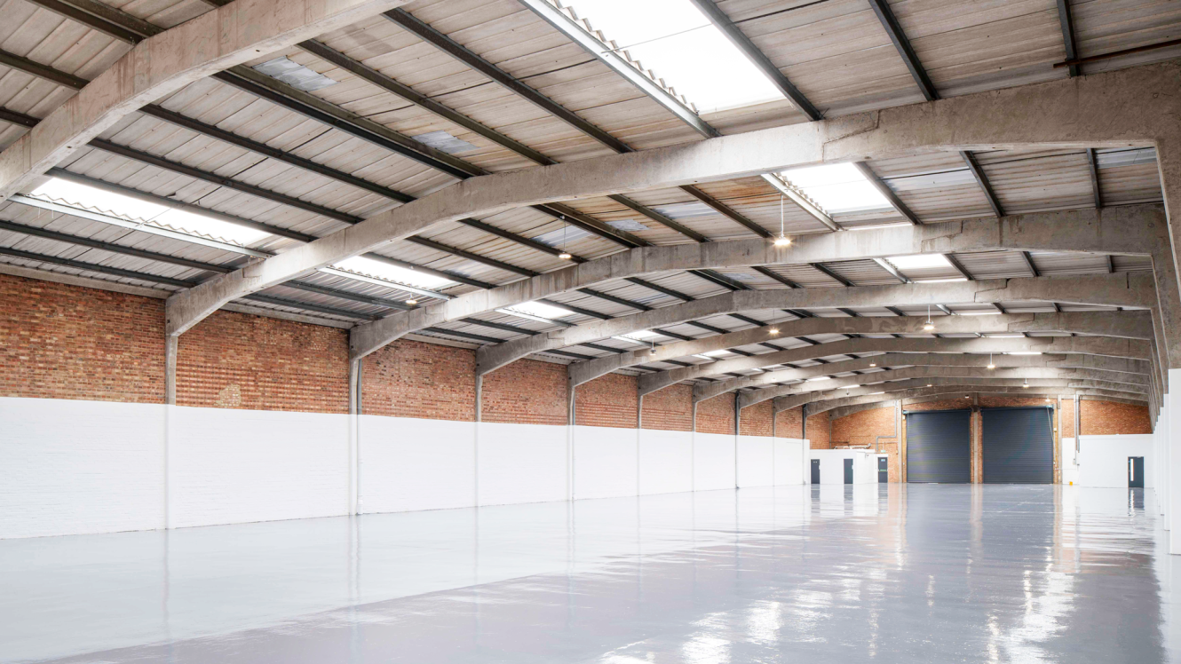 Large empty warehouse with brick walls, high ceiling, skylights, and a polished concrete floor. Several doors and a large black loading bay are visible along the back wall.