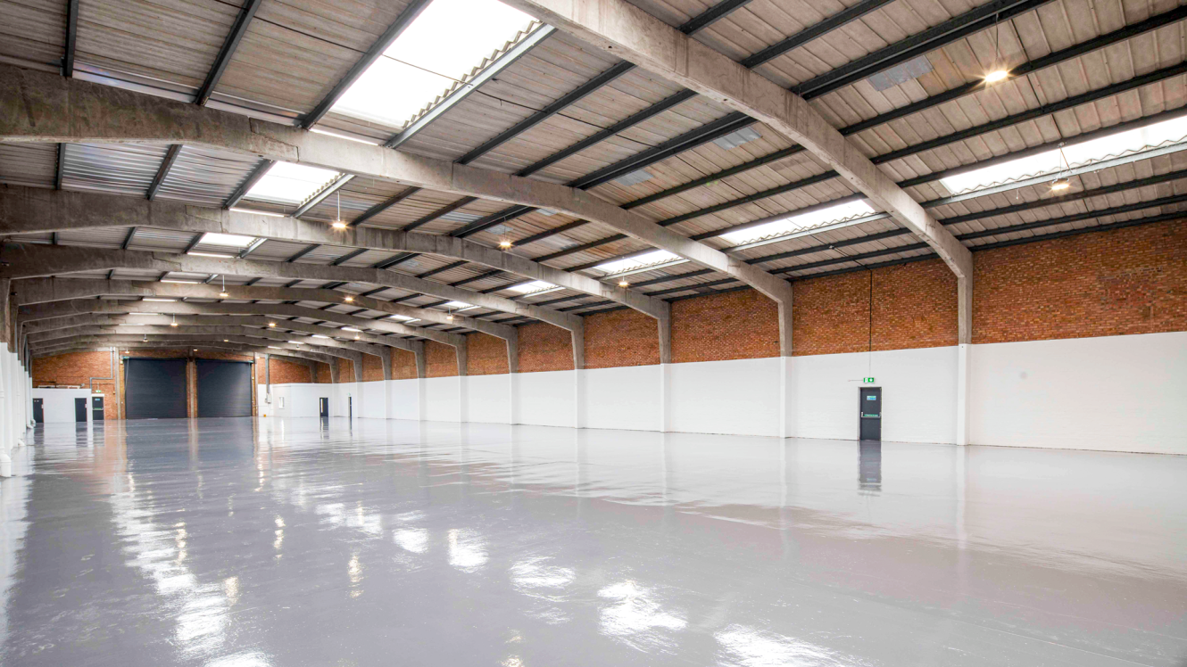 Large empty warehouse with a polished concrete floor, brick walls, high ceiling, skylights, and multiple doors along one wall.