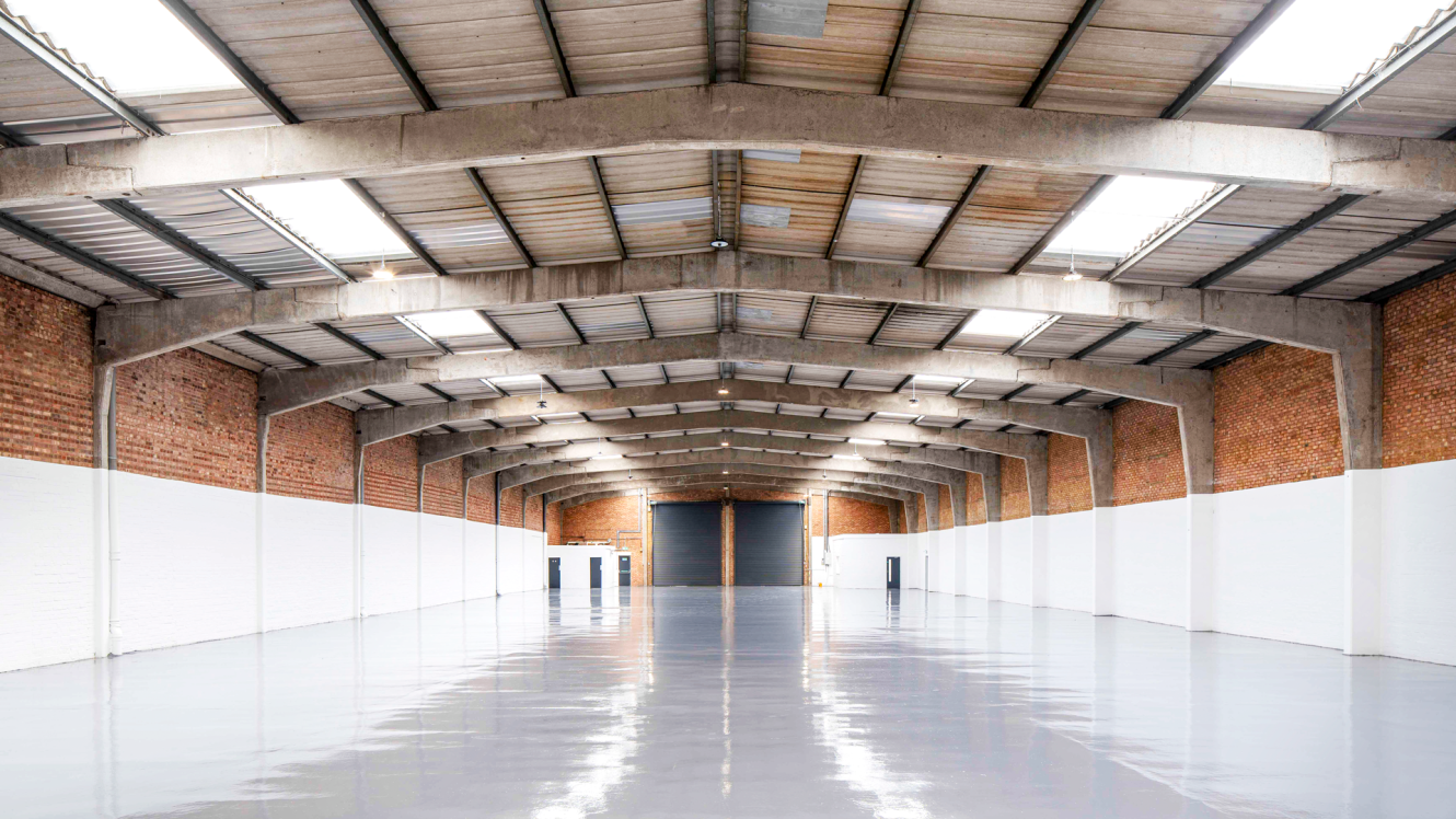 Empty warehouse with polished grey floors, exposed brick walls, and large skylights in the ceiling. A large closed door is visible at the far end of the space.