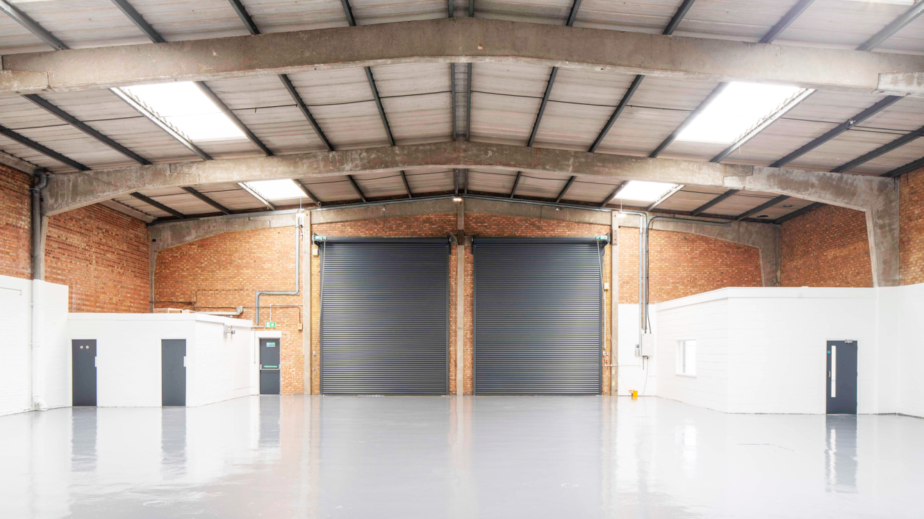 Interior of an empty warehouse with two large shutter doors, brick walls, a concrete ceiling, and two small white office rooms on either side.