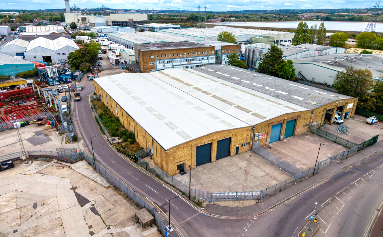 A large industrial warehouse with a white roof is surrounded by a fenced yard and adjacent roads in an industrial estate.