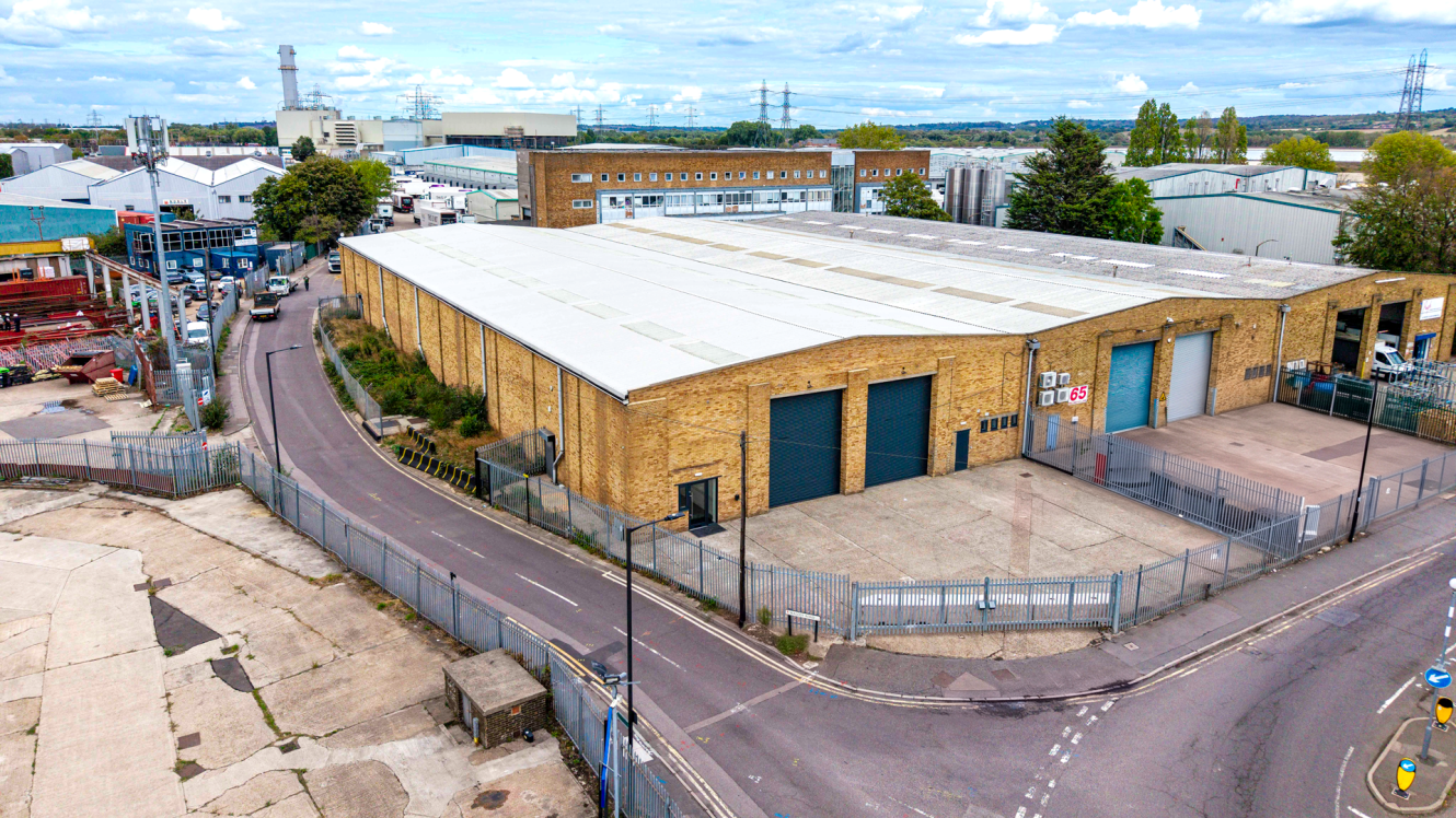 A large industrial warehouse with brick walls and multiple loading bays stands on a fenced yard in an industrial area surrounded by factories.