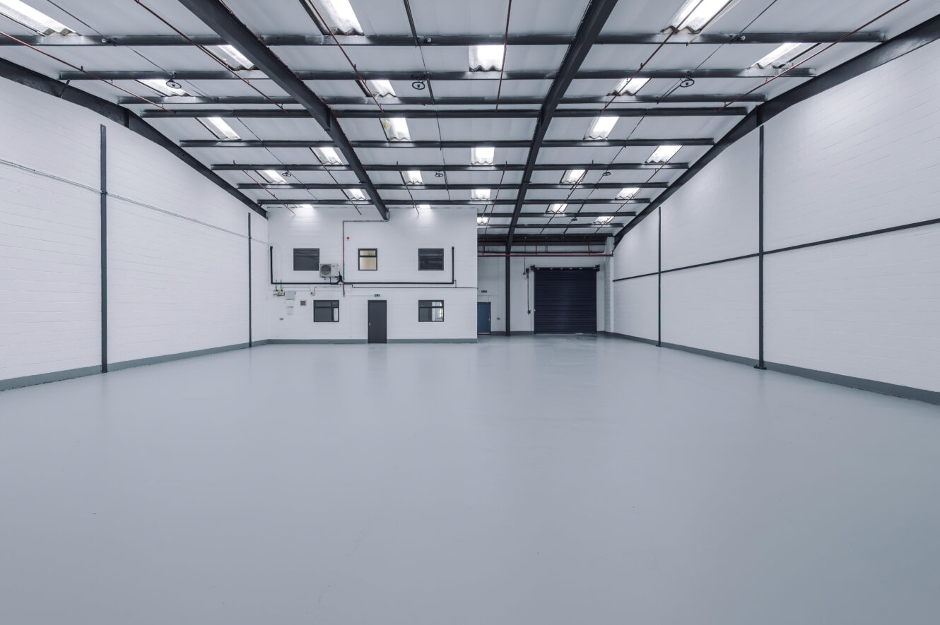 Interior of a large empty industrial warehouse with white walls, high ceilings, and fluorescent lights, featuring a small office section and a large black roller shutter door.
