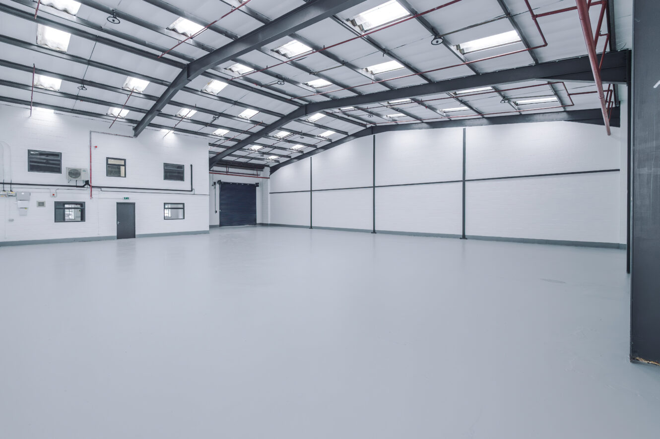 Empty industrial warehouse with white walls, high ceiling, exposed beams, windows, and a large roller shutter door, illuminated by ceiling lights.