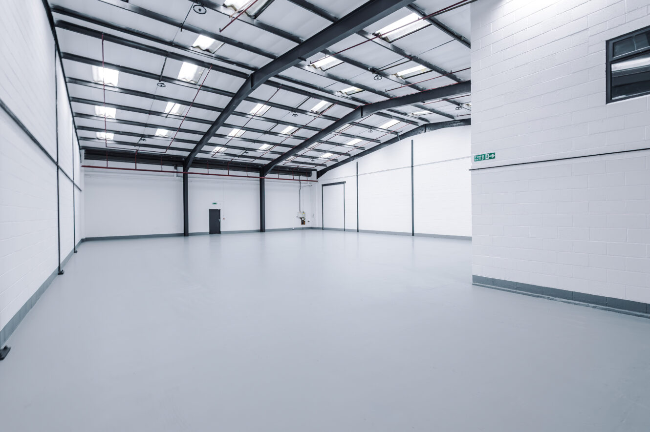Empty, clean industrial warehouse with white walls, a grey floor, high ceilings, exposed beams, and fluorescent lighting. An exit sign and doors are visible on the far side.